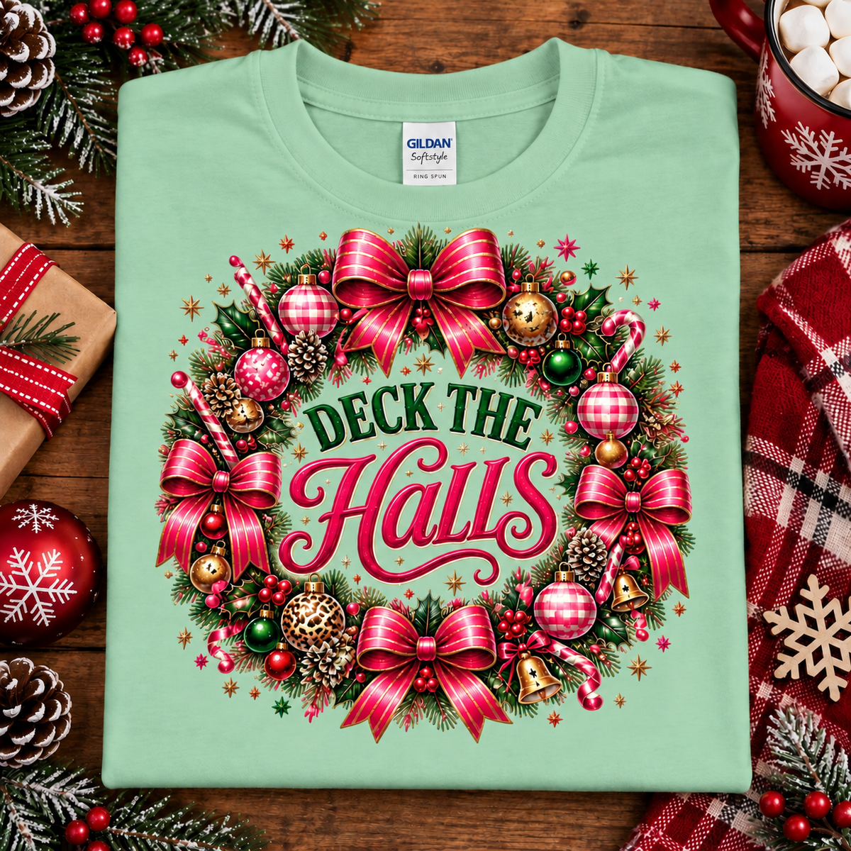 A folded mint green Gildan Signature T-shirt with a large festive Christmas wreath graphic and the words 'Deck the Halls' in red and green, styled on a wooden surface with holiday decorations.