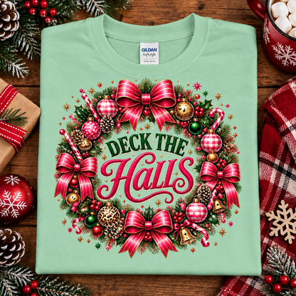 A folded mint green Gildan Signature T-shirt with a large festive Christmas wreath graphic and the words 'Deck the Halls' in red and green, styled on a wooden surface with holiday decorations.