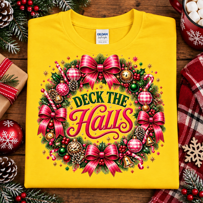 A folded bright yellow Gildan Softstyle t-shirt with a colorful Christmas wreath graphic reading 'Deck the Halls,' styled on a wooden surface with holiday ornaments, pinecones, wrapped gift boxes, and a red plaid scarf.