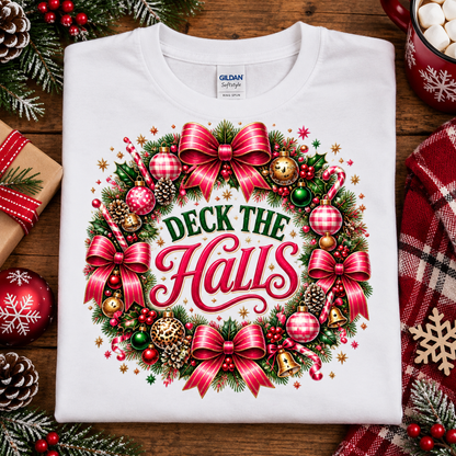 A folded white Gildan Comfort T-shirt with a colorful Christmas wreath graphic and the words 'DECK THE HALL' on the front, styled on a wooden surface with holiday decorations including pinecones, ornaments, wrapped gifts, and a red plaid cloth.