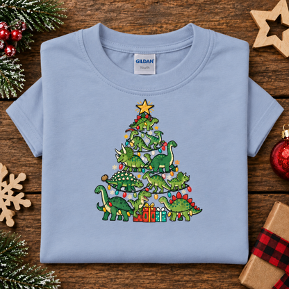 A light blue Gildan youth t-shirt folded flat on a rustic wooden background, decorated with Christmas tree graphics made of green dinosaurs, colorful ornaments, and presents. Styled with holiday props like pine branches, wooden stars, and a red ornament.