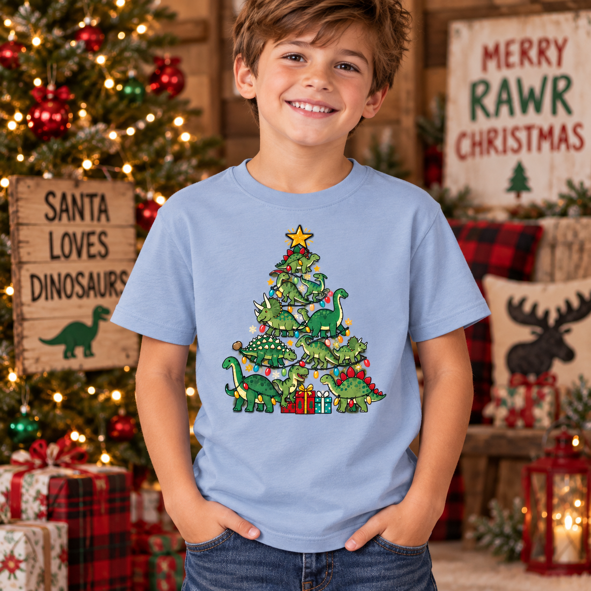 A smiling young boy wearing a light blue t-shirt with a green dinosaur Christmas tree graphic, standing in a cozy, decorated holiday room with Christmas lights and signs.
