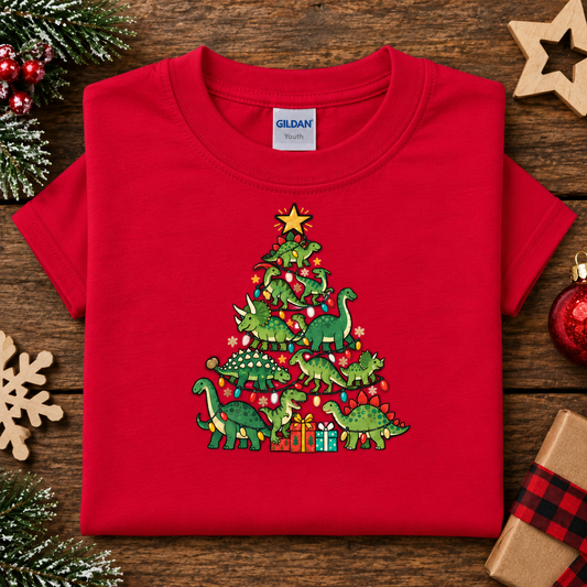 A folded red Gildan t-shirt with a Christmas tree made of green dinosaurs and red ornaments, displayed on a rustic wooden background with holiday decorations.