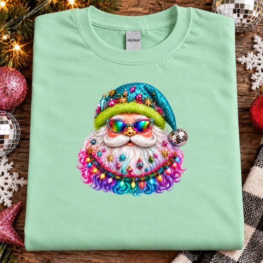 A mint green folded Gildan t-shirt with a colorful graphic of a Santa Claus face adorned with a sequined hat, rainbow sunglasses, and a fluffy beard decorated with Christmas ornaments. The shirt is displayed on a rustic wooden background surrounded by holiday decorations like a disco ball, snowflakes, and lights.