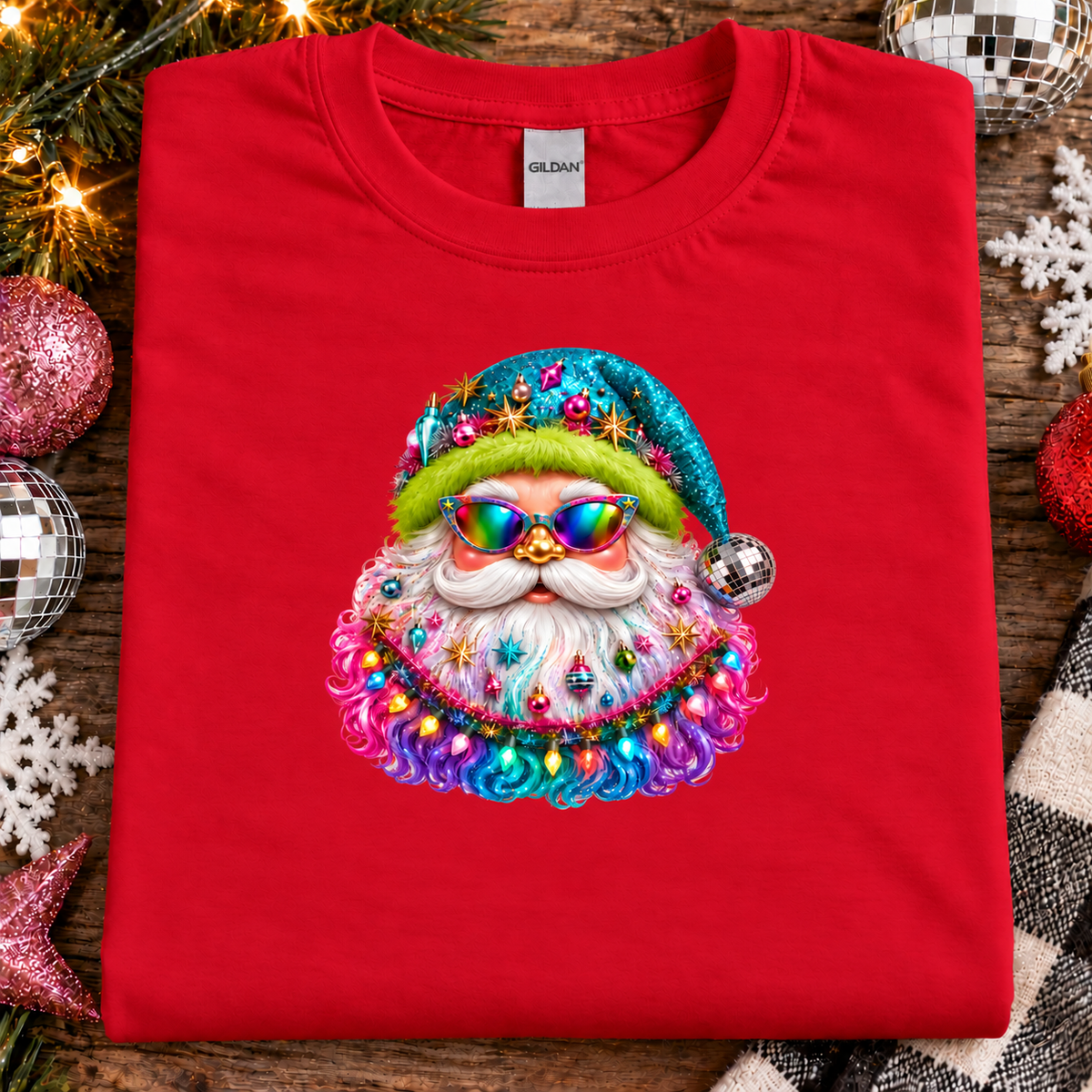 A folded bright red Gildan t-shirt featuring a colorful graphic of a Santa Claus face wearing a glittery blue hat, green hair, and rainbow sunglasses, styled on a rustic wooden background with holiday decorations.