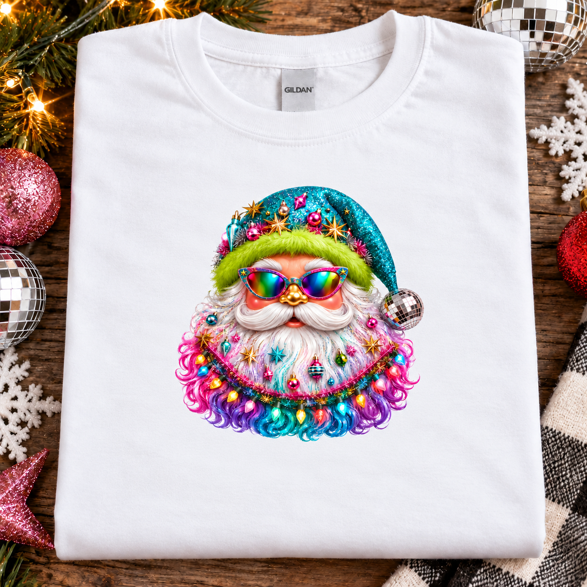A folded white Gildan t-shirt with a colorful, glittery Santa Claus graphic on the front, styled on a rustic wooden background with Christmas ornaments and lights.