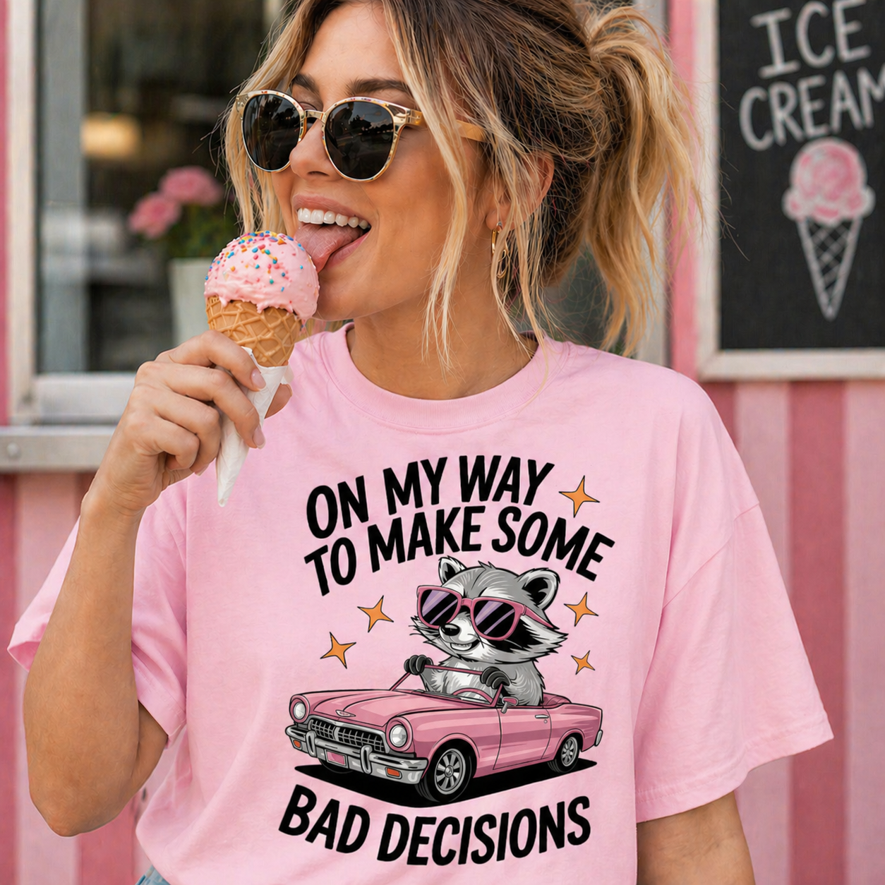 Woman wearing a blossom colored t-shirt with a raccoon graphic and text, standing in front of an ice cream shop.