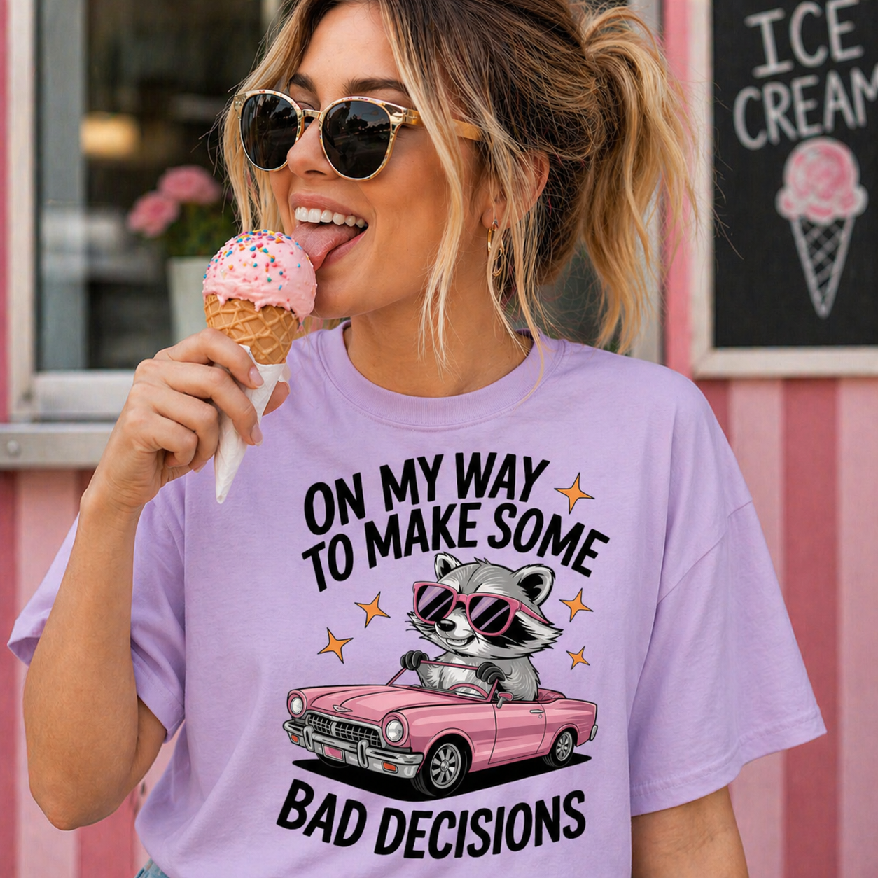 Woman wearing an orchid colored t-shirt with a raccoon graphic and text, eating ice cream in front of an ice cream shop.