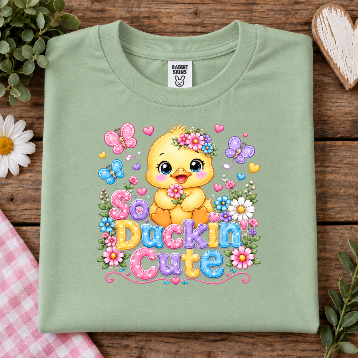 A folded sage green t-shirt with a colorful graphic of a yellow duckling surrounded by flowers and the text 'So Duckin Cute'. The shirt is displayed on a rustic wooden background with decorative props including a daisy, green leaves, a heart-shaped wooden ornament, and a pink gingham cloth.