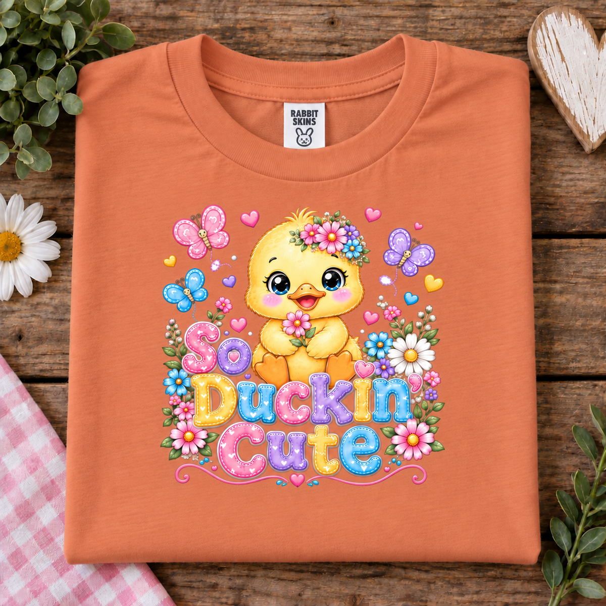 An sunset orange crewneck t-shirt by Rabbit Skin's, featuring a colorful graphic of a yellow duckling surrounded by flowers, butterflies, and hearts with the text 'So Duckin Cute'. The shirt is displayed in a flat-lay arrangement on a rustic wooden background with decorative elements like a daisy, green leaves, and a heart-shaped wooden ornament.