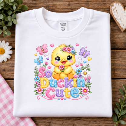 A folded white t-shirt with a colorful graphic of a cute yellow duckling surrounded by flowers, butterflies, and the text 'So Duckin Cute'. The shirt is displayed flat on a rustic wooden background with decorative props including a pink gingham cloth, a white daisy, and a wooden heart.