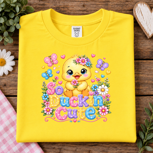 A folded bright yellow t-shirt with a colorful graphic of a cute yellow duckling surrounded by flowers, butterflies, and hearts, with the text 'So Duckin Cute'. The shirt is displayed on a rustic wooden background with decorative spring elements like a daisy, eucalyptus leaves, and a heart-shaped wooden ornament.