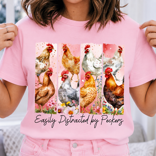 Blossom colored t-shirt with chicken design and text 'Easily Distracted by Peckers' held by a person.