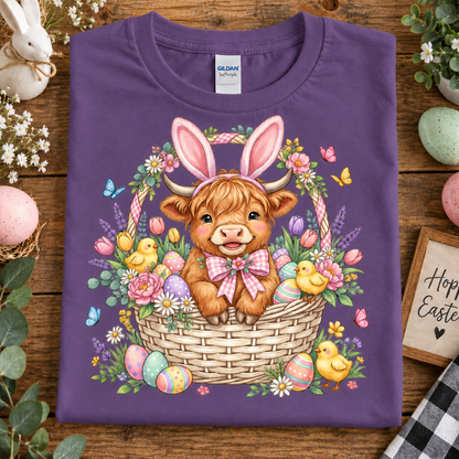 A folded Heather purple Gildan Softstyle T-shirt with a colorful Easter graphic of a Scottish Highland cow in a basket, surrounded by chicks, flowers, and eggs, displayed on a wooden surface with decorative Easter props.