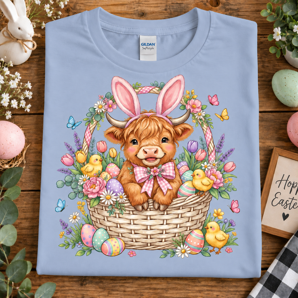 A folded light blue Gildan Softstyle T-shirt with a colorful Easter graphic of a Scottish highland cow in a basket with flowers, chicks, and eggs, styled on a wooden surface with Easter props.