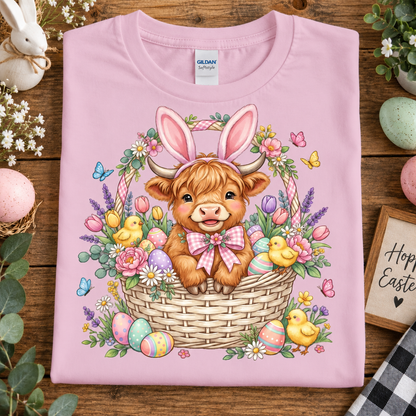 A folded light pink Gildan t-shirt with a colorful Easter graphic of a Scottish Highland cow in a basket, surrounded by flowers, chicks, and Easter eggs, displayed on a rustic wooden background with decorative props.