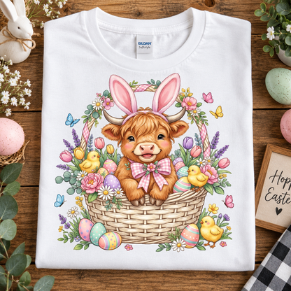 A folded white Gildan Softstyle T-shirt with a colorful Easter graphic of a Scottish highland cow in a basket, surrounded by flowers, chicks, and pastel eggs, displayed on a wooden surface with Easter-themed props.