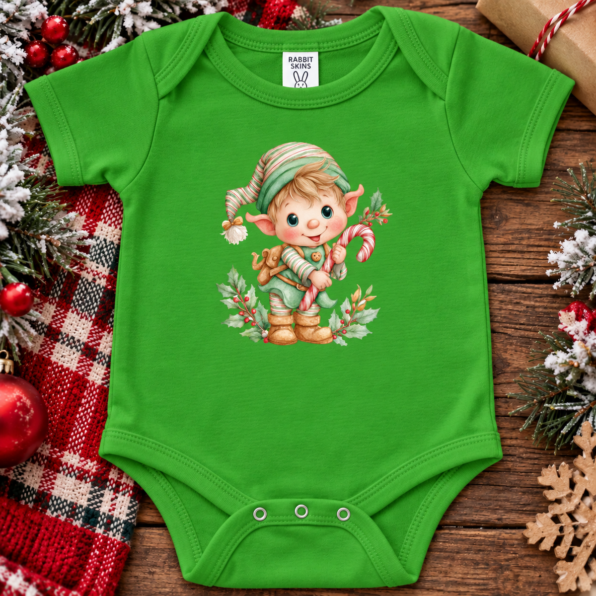 A apple green baby bodysuit with a colorful Christmas elf graphic, laid flat on a rustic wooden surface surrounded by holiday decorations including a red plaid blanket, pine branches, ornaments, and wrapped gifts. The brand label inside the neck reads Rabbit Rich.