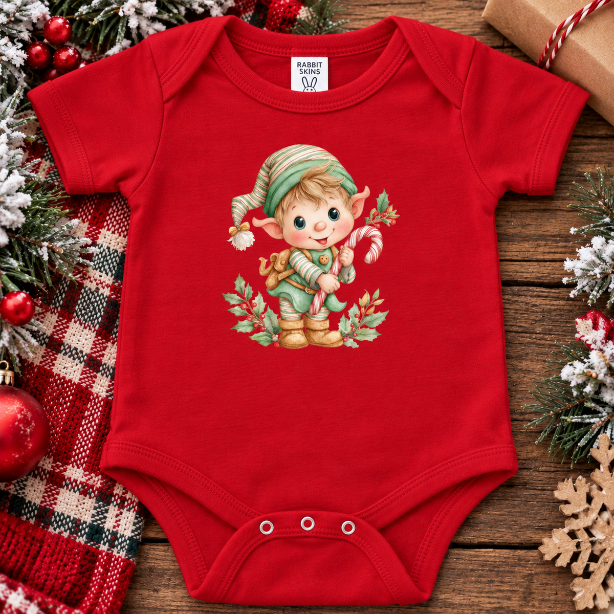 A red baby bodysuit with a festive Christmas elf graphic, styled in a flat-lay on a rustic wooden background with holiday decorations including a plaid blanket, ornaments, and pine branches.