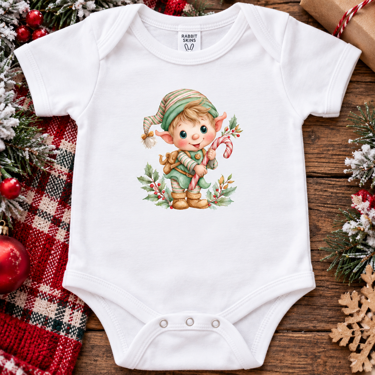 A white short-sleeve baby bodysuit with a colorful Christmas elf graphic on the chest, styled in a flat lay on a rustic wooden surface with holiday decorations including a red plaid blanket, pine branches, ornaments, and a snowflake.