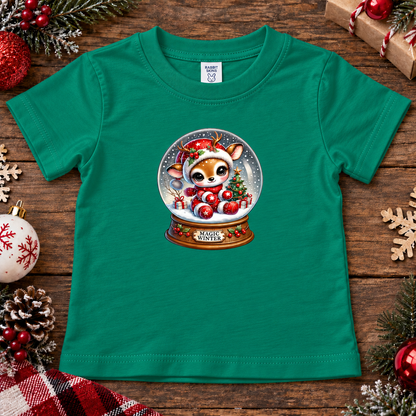 A green short-sleeve baby t-shirt laid flat on a rustic wooden surface, decorated with Christmas ornaments, pinecones, and evergreen branches. The center of the shirt features a graphic of a cute cartoon puppy inside a snow globe with red ornaments and the text 'MERRY CHRIST'.