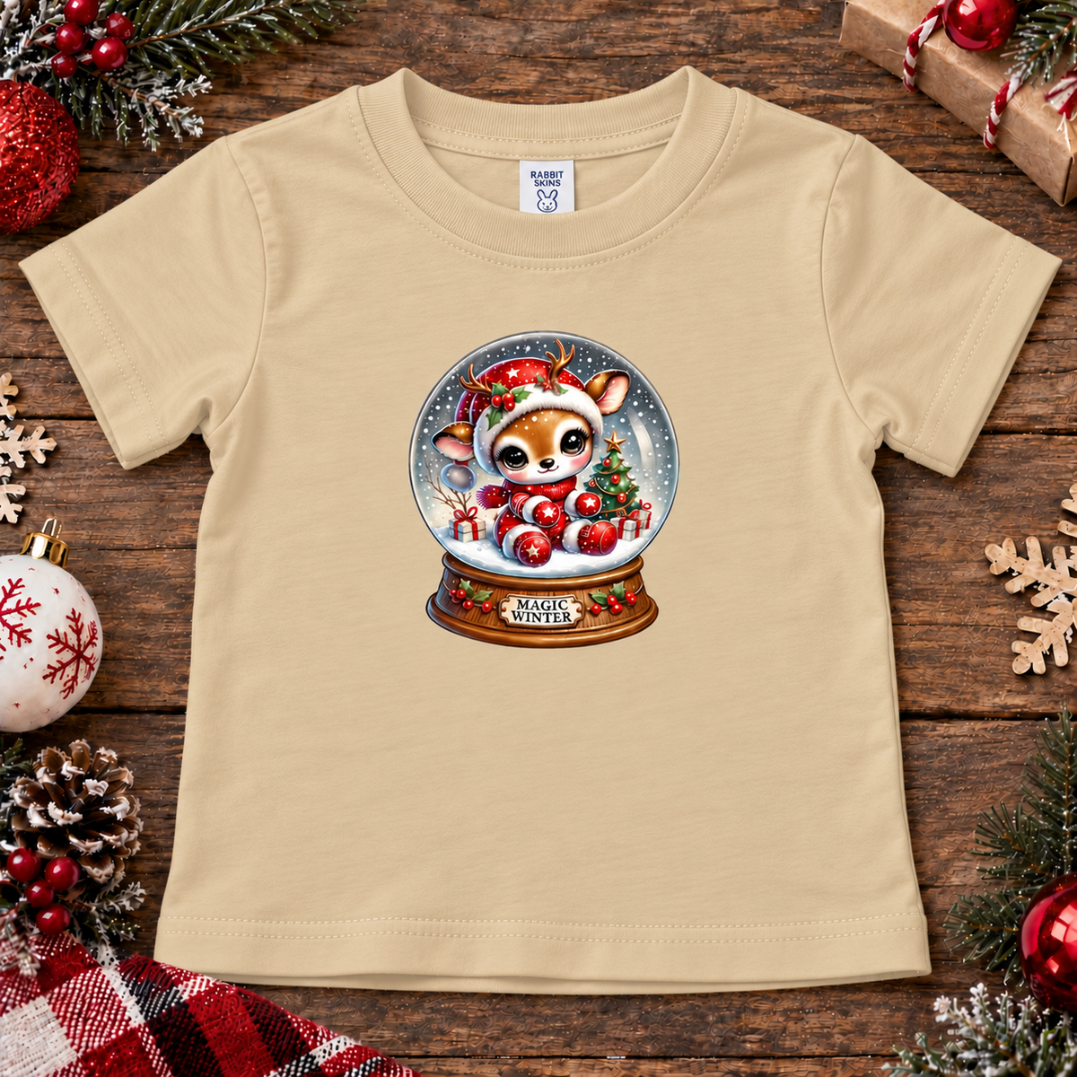 A latte toddler short-sleeve t-shirt with a Christmas snow globe graphic featuring a cute deer and ornaments, laid flat on a rustic wooden background decorated with holiday ornaments, pinecones, and a gift box.