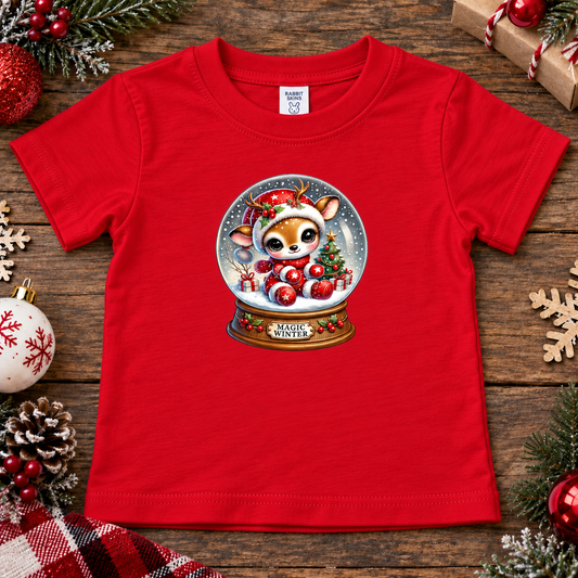 A red short-sleeve kids' t-shirt with a Christmas-themed graphic of a cartoon deer inside a snow globe. The shirt is displayed in a flat-lay setting on a rustic wooden background, surrounded by holiday decorations like pine branches, ornaments, and a plaid blanket.