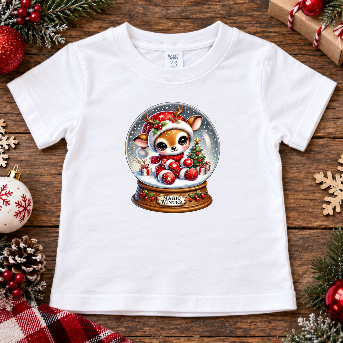 A white short-sleeve baby t-shirt laid flat on a rustic wooden background, decorated with Christmas ornaments, pine branches, and a wrapped gift. The front of the shirt features a colorful graphic of a cute deer inside a 'Magic Winter' snow globe.