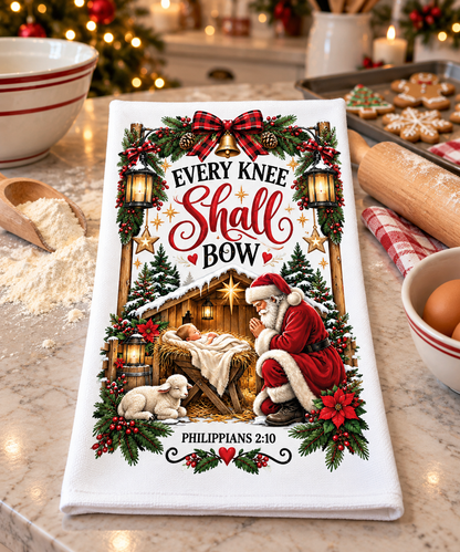 A white kitchen towel with a detailed Christmas nativity print featuring Santa Claus, a baby in a manger, a lamb, and the text 'EVERY KNEE Shall BOW' and 'PHILIPPIANS 2:10', styled on a marble countertop with baking props.