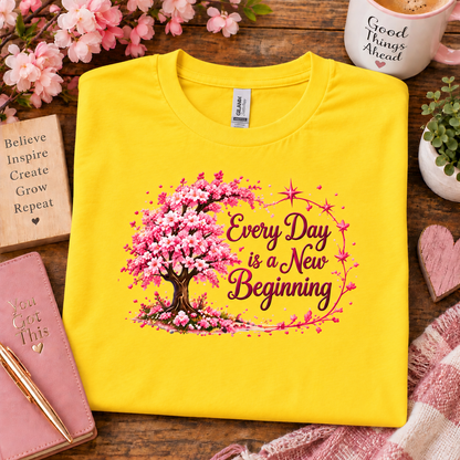 A folded daisy yellow T-shirt with a cherry blossom tree graphic and the phrase 'Every Day is a New Beginning' in pink and brown lettering, styled on a wooden surface with flowers, a mug, a notebook, and a scarf.