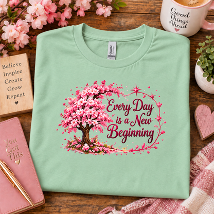 A mint green crewneck sweatshirt with a cherry blossom tree graphic and the text 'Every Day is a New Beginning' laid flat on a wooden table surrounded by lifestyle props including a notebook, flowers, a mug, and a scarf.