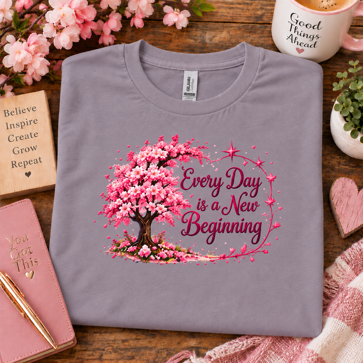 A folded paragon purple-gray t-shirt with a cherry blossom tree graphic and the text 'Every Day is a New Beginning' in pink and burgundy, displayed on a wooden desk with a notebook, pen, mug, and plants.