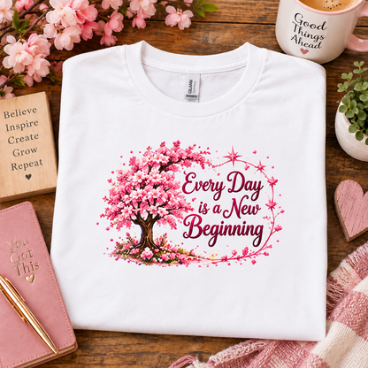 A white crewneck sweatshirt with a cherry blossom tree graphic and the text 'Every Day is a New Beginning' laid flat on a wooden table surrounded by decorative props including a notebook, flowers, a mug, and a plant.