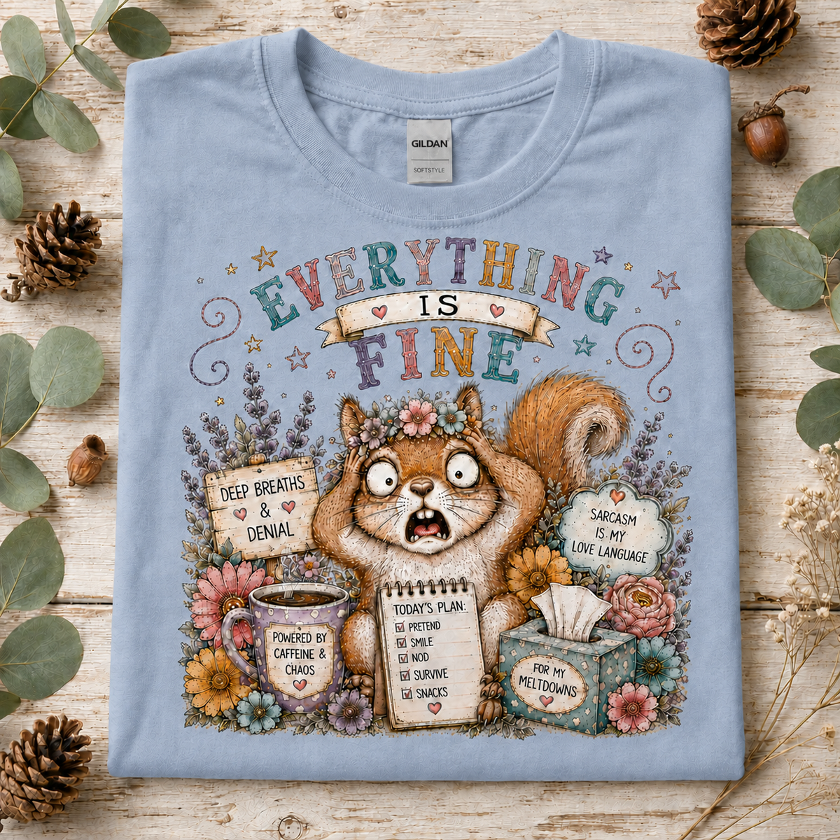 A light blue graphic t-shirt laid flat on a rustic wooden surface surrounded by eucalyptus leaves and pinecones. The shirt features a colorful cartoon illustration of a squirrel surrounded by flowers and humorous text bubbles.