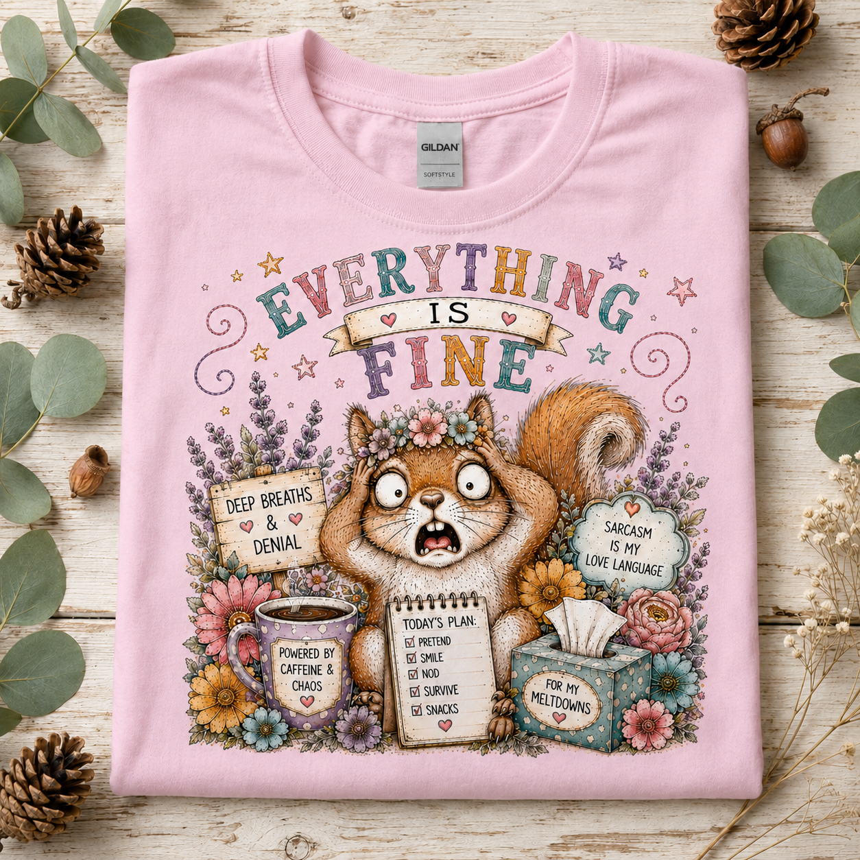 A light pink short-sleeve t-shirt with a detailed graphic print of a squirrel surrounded by flowers and humorous text, laid flat on a rustic wooden surface with eucalyptus leaves and pinecones.