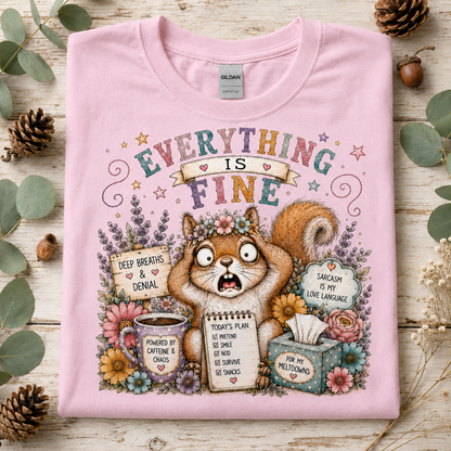 A light pink short-sleeve t-shirt with a detailed graphic print of a squirrel surrounded by flowers and humorous text, laid flat on a rustic wooden surface with eucalyptus leaves and pinecones.
