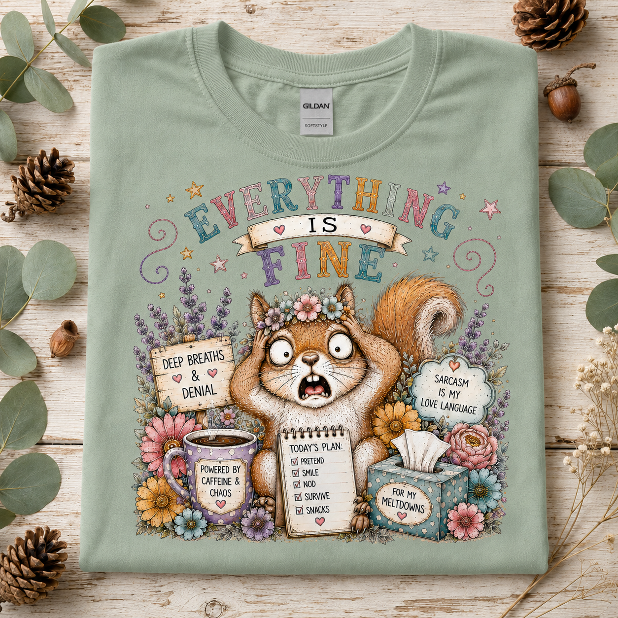 A sage green crewneck t-shirt with a colorful cartoon squirrel graphic and text reading 'EVERYTHING IS FINE', laid flat on a rustic wood surface surrounded by eucalyptus leaves and pinecones.