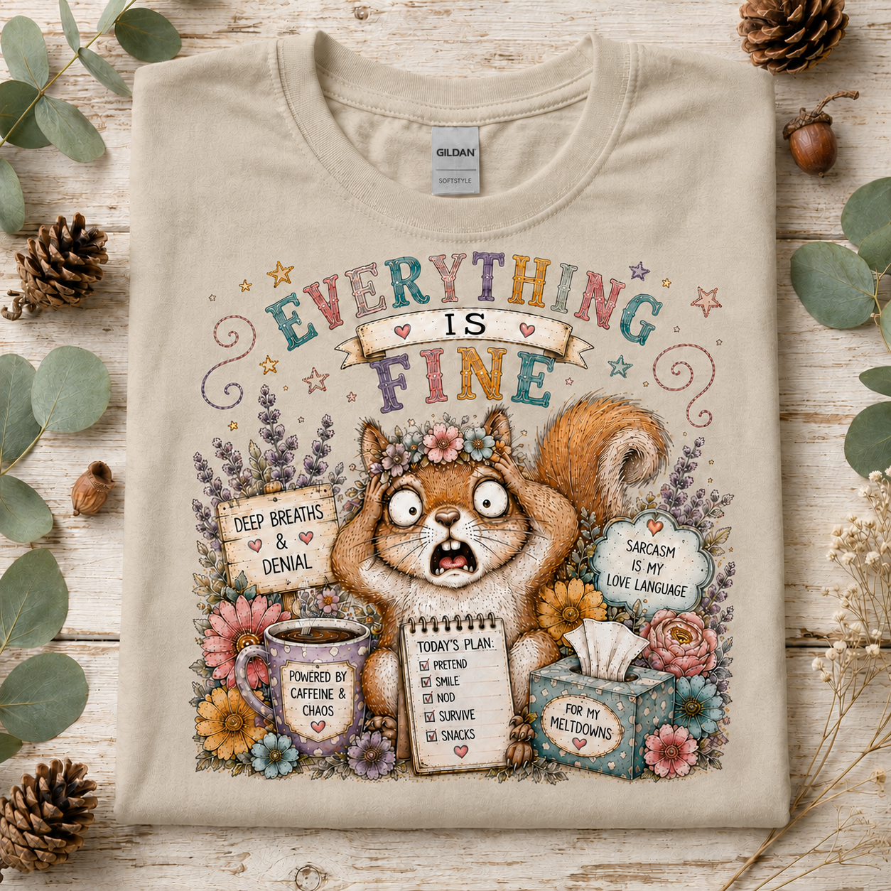A sand colored graphic T-shirt laid flat on a rustic wood surface with eucalyptus leaves and pinecones. The shirt features a colorful illustrated squirrel surrounded by flowers and text that reads “EVERYTHING IS FINE,” with additional humorous labels and drawings.
