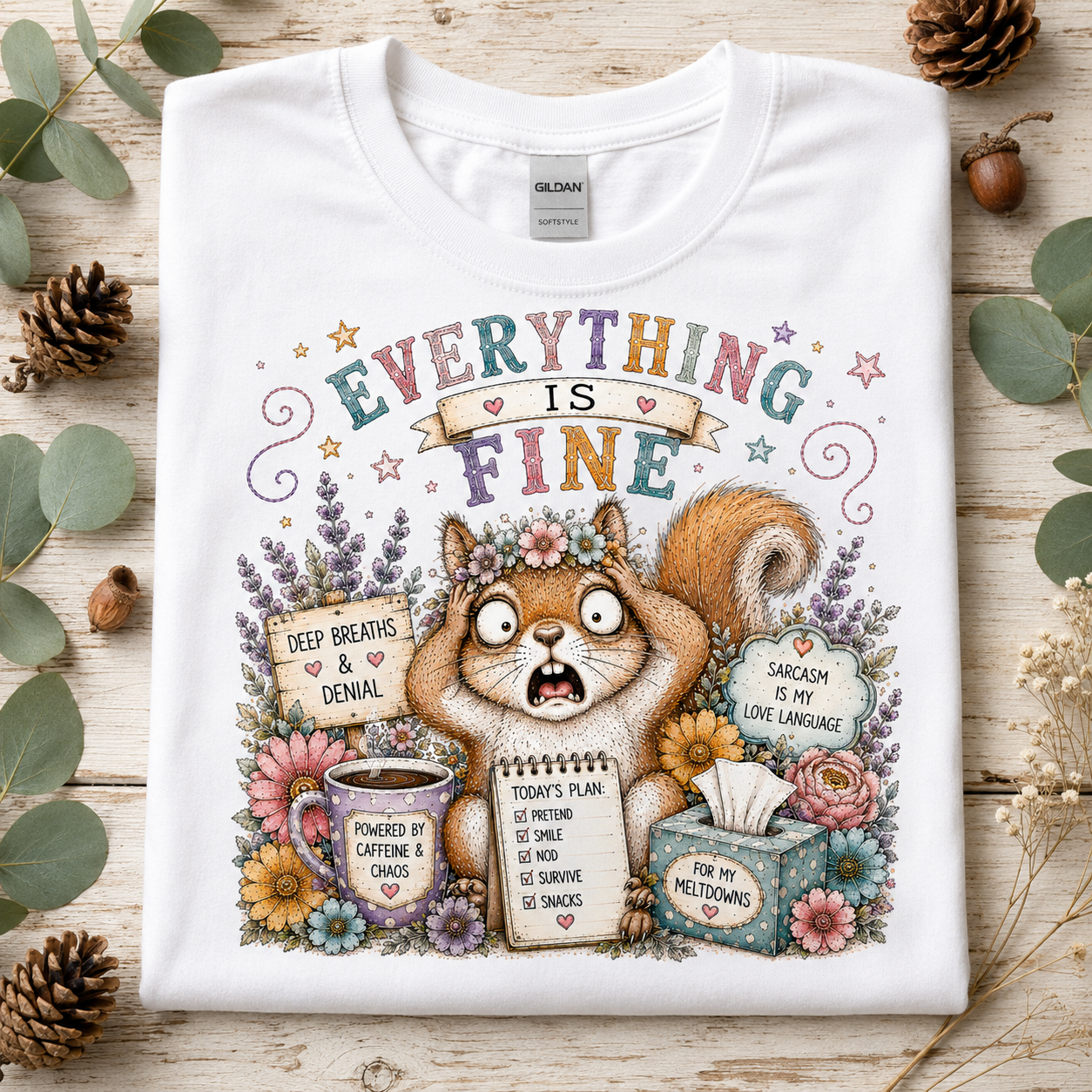 A white Gildan t-shirt laid flat on a rustic wooden surface, surrounded by decorative green leaves, pinecones, and acorns. The shirt features a colorful graphic of a surprised squirrel surrounded by flowers and text that reads 'EVERYTHING IS FINE' with humorous coping labels.