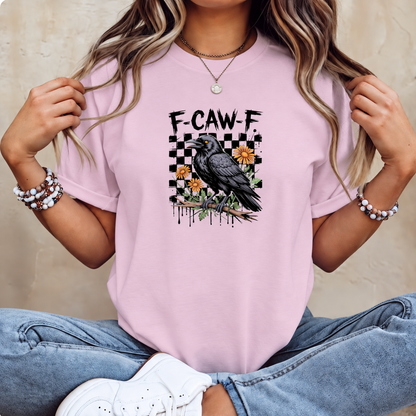 A woman wearing a light pink graphic t-shirt with a black raven and the text 'F-CAW-F' on the front, paired with blue jeans and white sneakers.