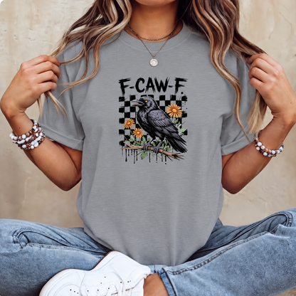 A woman wearing a gray graphic t-shirt with a black raven, yellow flowers, and the text 'F-CAW-F' on the front, paired with blue jeans and white sneakers.