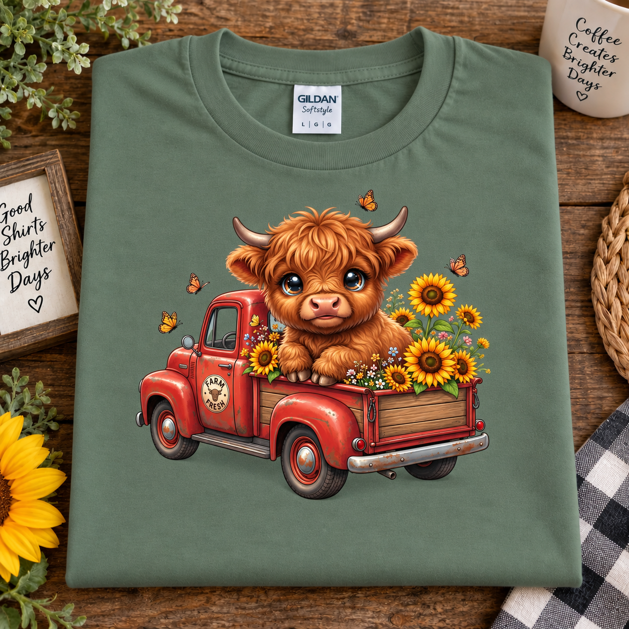 An military green Gildan Straightforward t-shirt is laid flat on a rustic wooden table, featuring a cute graphic of a fluffy brown Highland cow sitting in a red vintage truck filled with yellow sunflowers. The shirt is styled with decorative props including a sunflower, a mug, and a small sign.