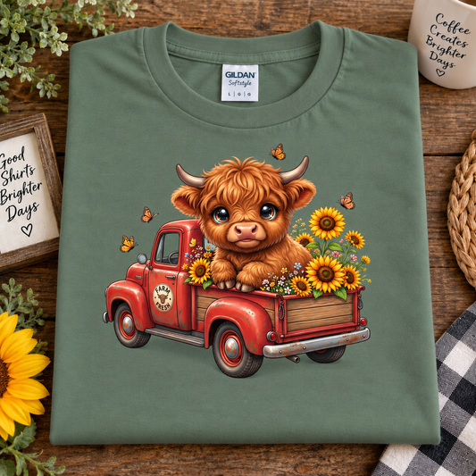 An military green Gildan Straightforward t-shirt is laid flat on a rustic wooden table, featuring a cute graphic of a fluffy brown Highland cow sitting in a red vintage truck filled with yellow sunflowers. The shirt is styled with decorative props including a sunflower, a mug, and a small sign.