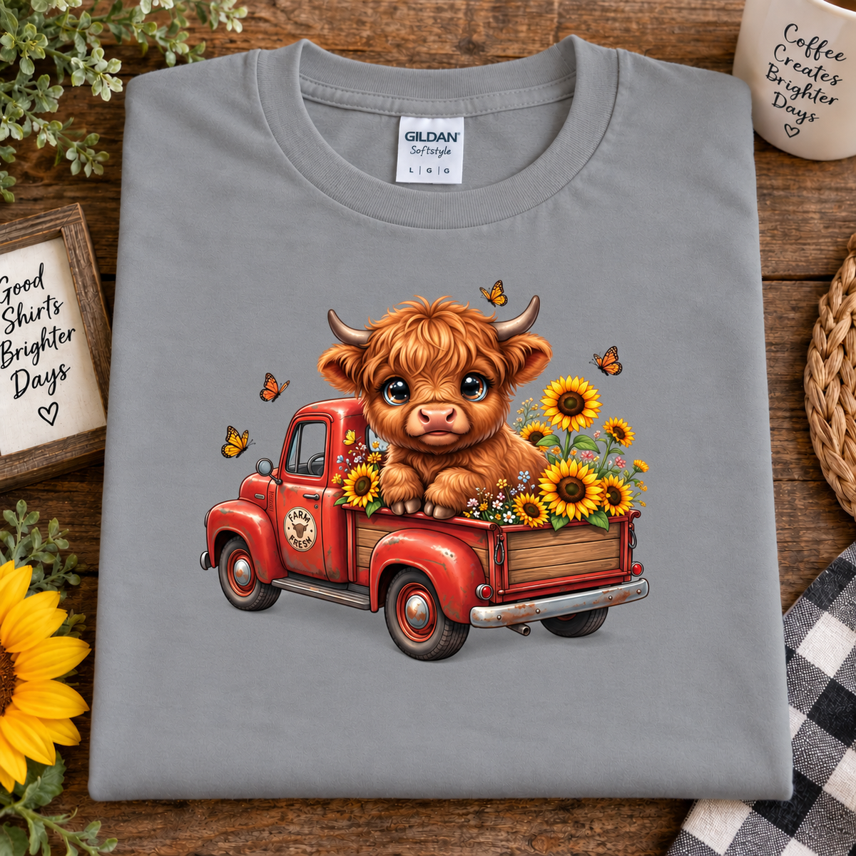 A folded sport gray Gildan Straightweight t-shirt is displayed on a rustic wooden background with decorative props like a sunflower, a mug, and a woven basket. The shirt features a colorful graphic of a cute brown Highland cow sitting in a red vintage pickup truck filled with yellow sunflowers and butterflies.