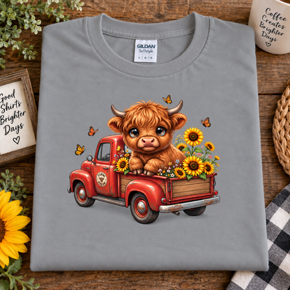 A folded sport gray Gildan Straightweight t-shirt is displayed on a rustic wooden background with decorative props like a sunflower, a mug, and a woven basket. The shirt features a colorful graphic of a cute brown Highland cow sitting in a red vintage pickup truck filled with yellow sunflowers and butterflies.
