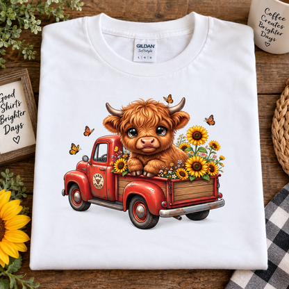 A folded white Gildan Straightforward t-shirt is displayed on a rustic wooden table. The shirt features a colorful graphic print of a cute, fluffy brown Scottish highland cow sitting in the bed of a red vintage pickup truck filled with yellow sunflowers and small butterflies.