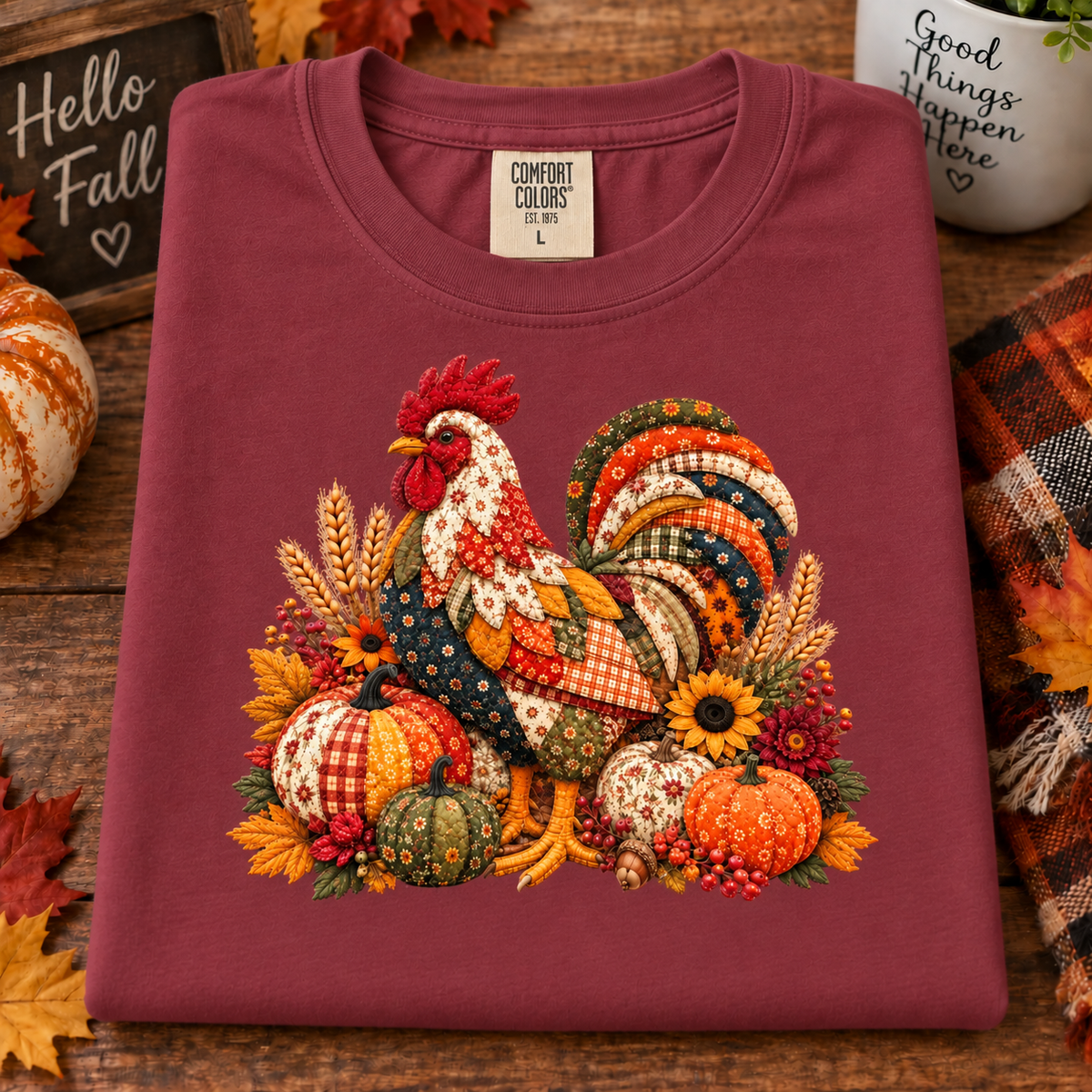 A folded chili colored t-shirt with a colorful autumn-themed rooster and pumpkins graphic, displayed on a rustic wooden surface with seasonal props.