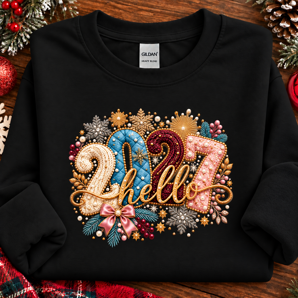 A black long-sleeve crewneck sweatshirt laid flat on a rustic wooden surface with holiday decorations around it. The front has a large decorative graphic reading “2027 hello” in glittery gold, blue, pink, and red tones, surrounded by faux beads, pinecones, berries, and winter motifs.