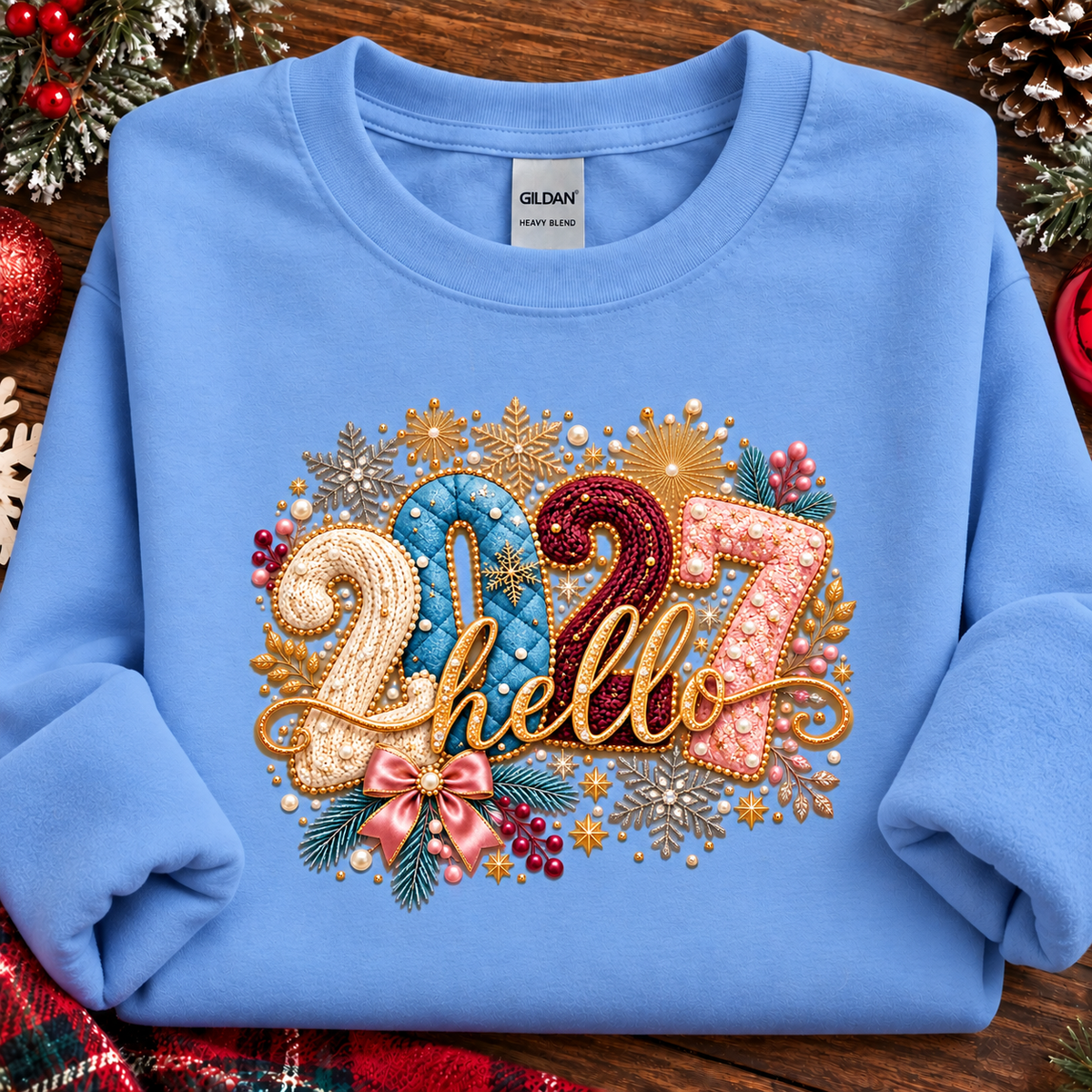 A folded carolina blue crewneck sweatshirt with a large, festive '2027 hello' graphic on the front. The graphic features gold glitter-style lettering and numbers embellished with sequins, beads, pinecones, and green pine needles.
