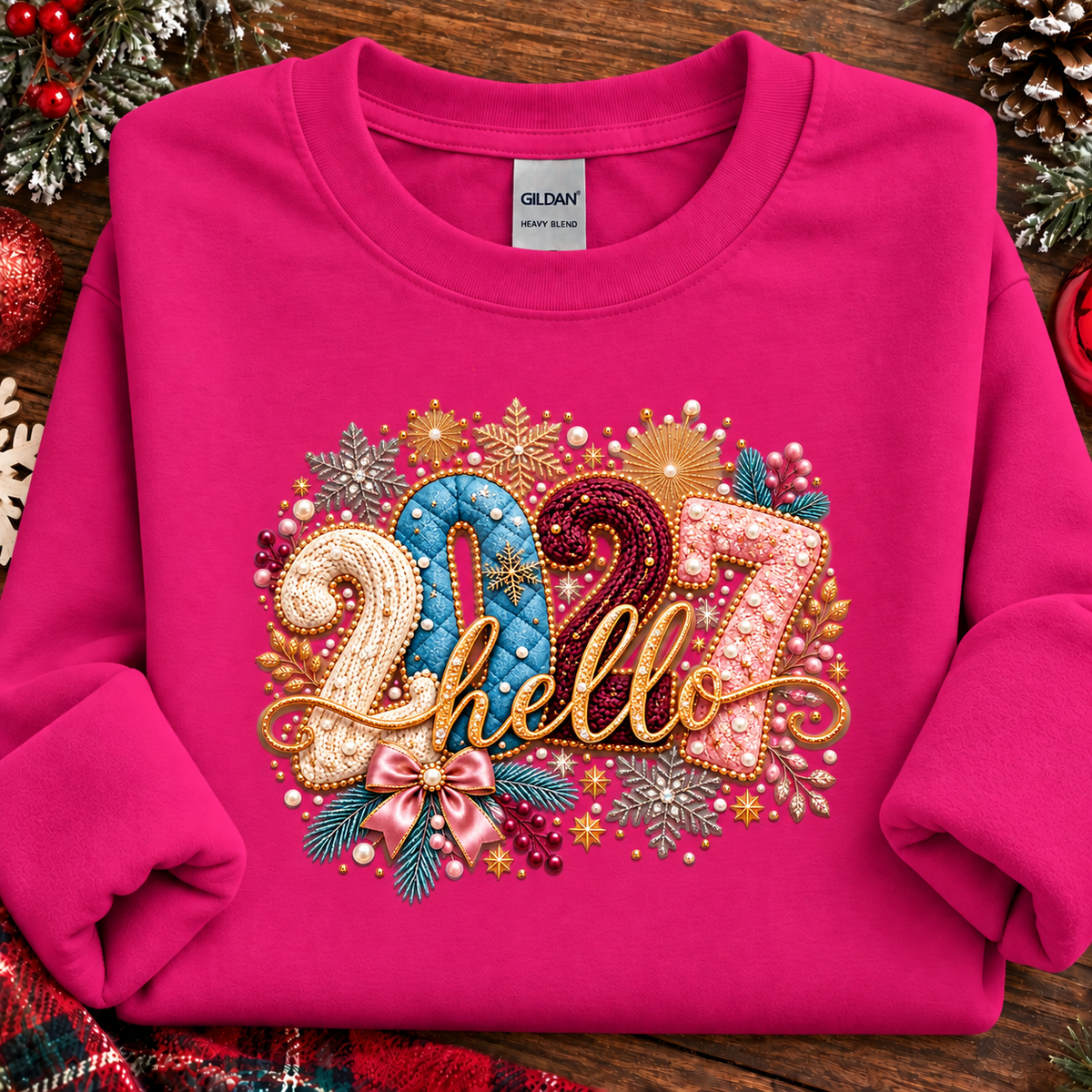 A folded heliconia pink sweatshirt on a rustic wooden surface with holiday decorations around it. The front has a large decorative graphic reading “2027 hello” in gold, blue, pink, and white embellished lettering with snowflakes, stars, and ribbon details.